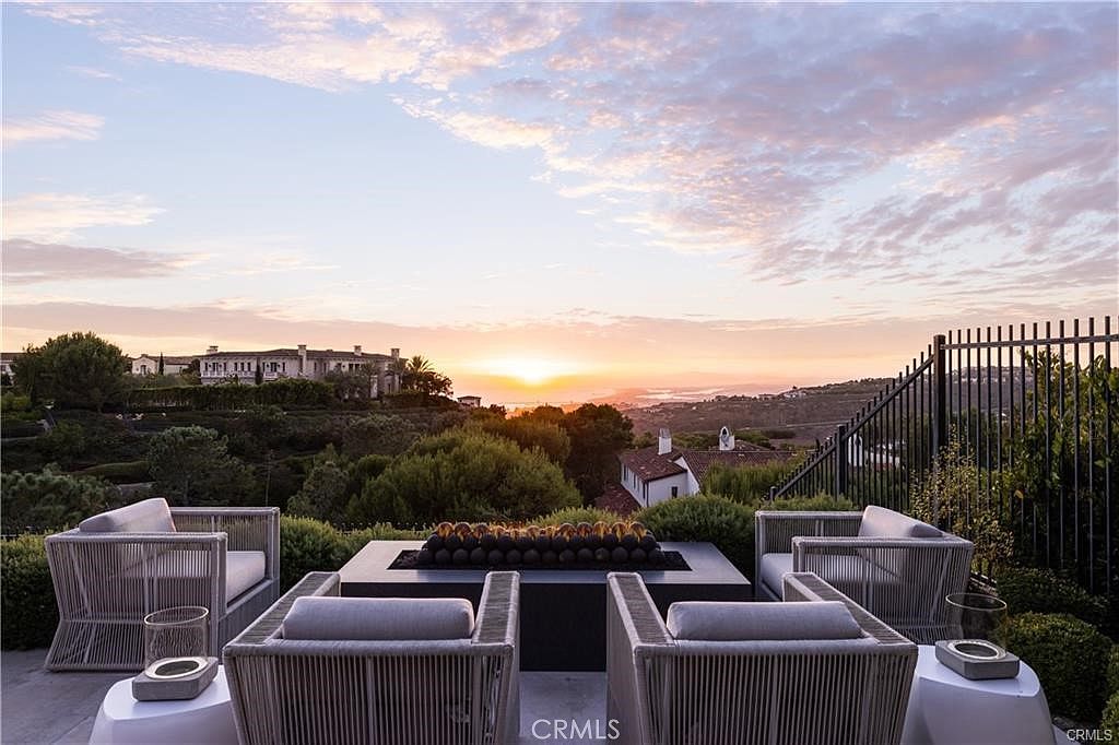 This elevated outdoor patio offers a stunning sunset view over a rolling landscape and distant coastline. The space is furnished with four modern, woven-style armchairs arranged around a central rectangular fire pit filled with dark stones. The scene is framed by a black metal railing, creating a sophisticated and serene atmosphere perfect for evening relaxation.