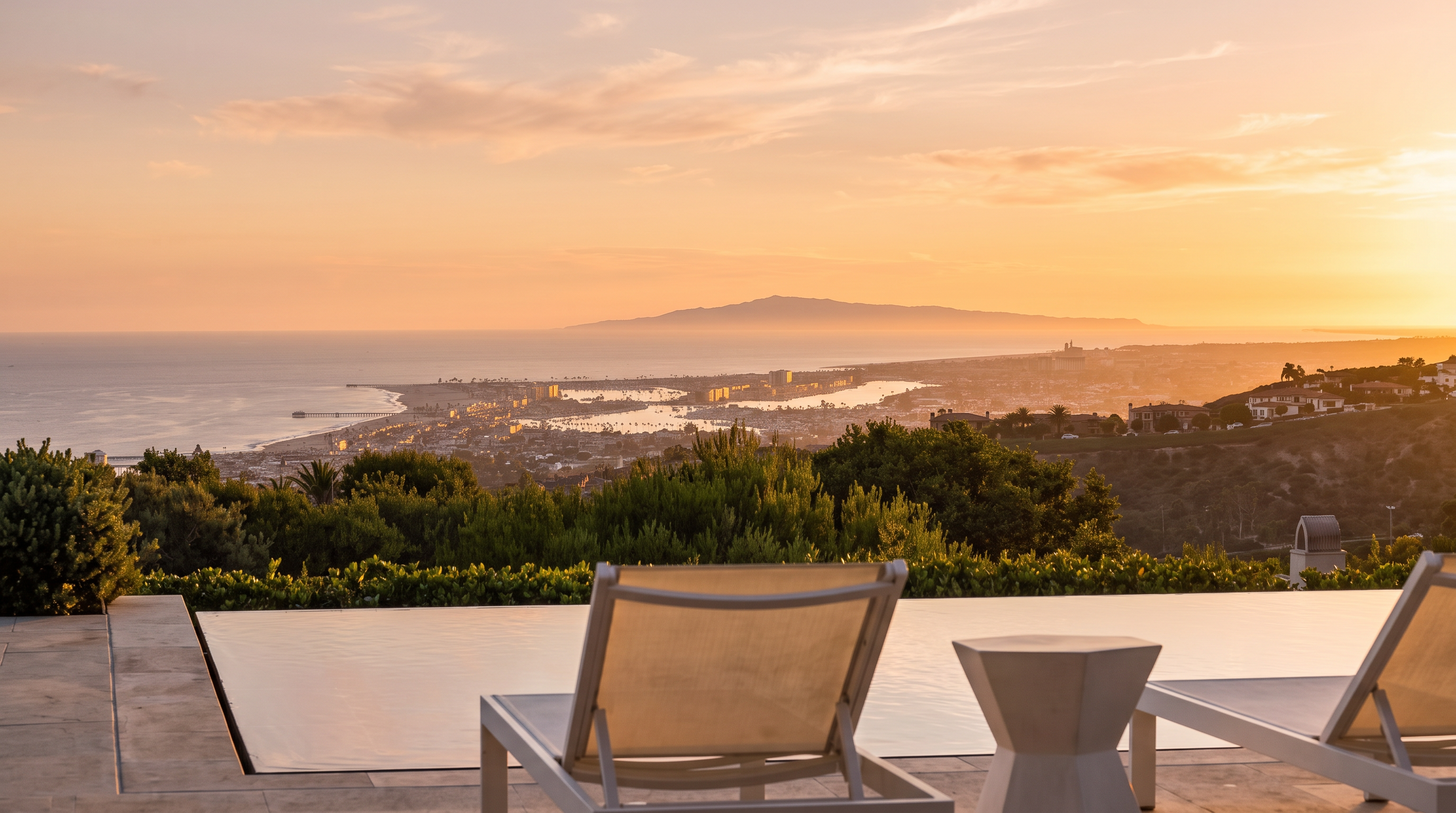 This image captures a stunning infinity-edge pool overlooking a vast coastal landscape, including a harbor, beach, and distant mountains. In the foreground, two white lounge chairs are positioned on a light-toned patio, inviting relaxation while taking in the expansive panoramic view. The perspective is set from a low angle, emphasizing the seamless transition between the pool water and the horizon beyond.