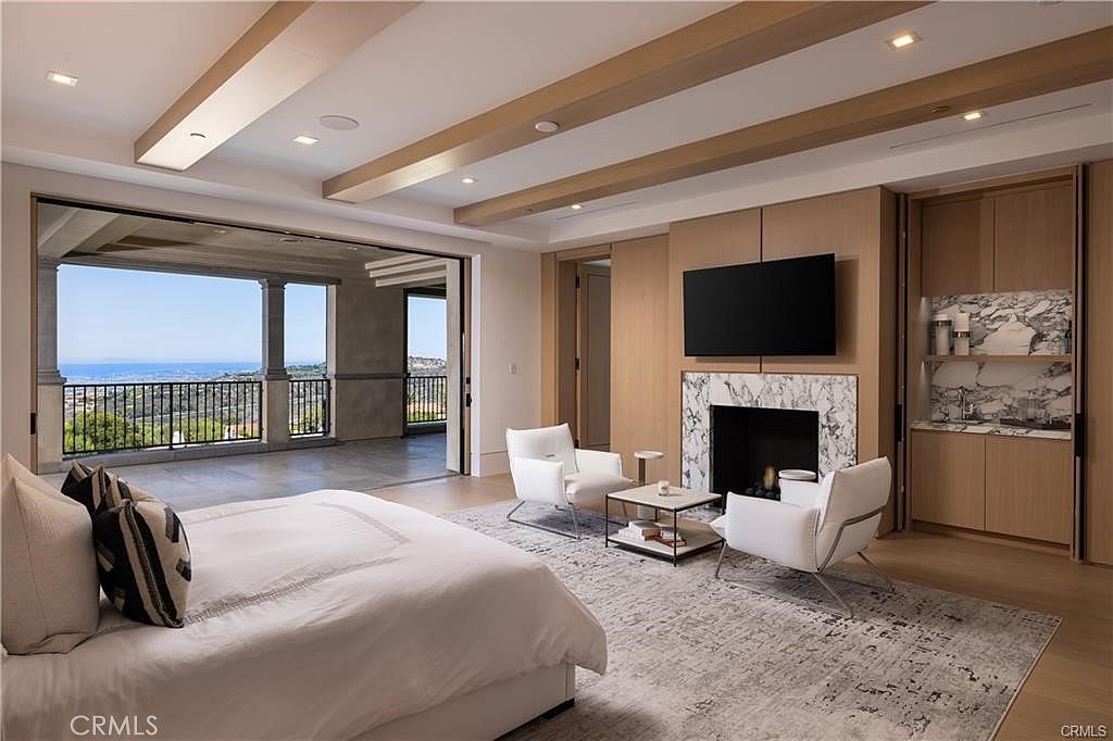 This expansive primary bedroom features a sophisticated, neutral palette with light wood paneling and a prominent stone-clad fireplace surround. A large bed sits in the foreground, while two white armchairs and a small table create a cozy seating area near the fireplace and a built-in wet bar. The room opens onto a spacious covered balcony through wide-spanning glass doors, offering a panoramic view of the coastline and hills.
