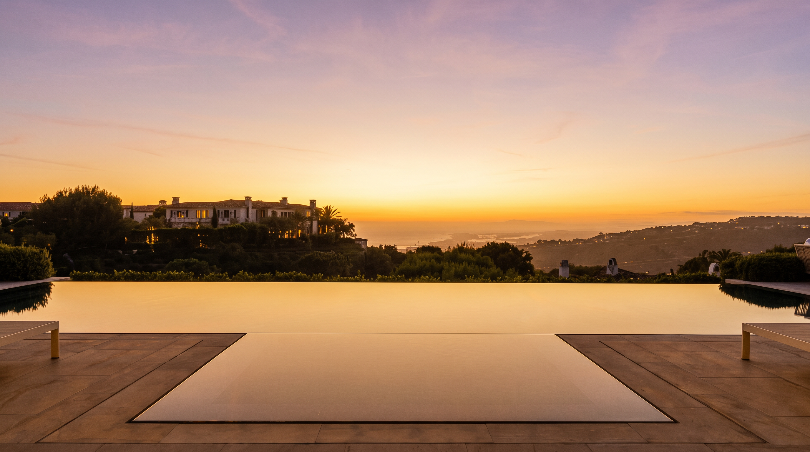 This image captures a stunning infinity-edge pool overlooking a vast, rolling landscape at sunset. The foreground features a paved deck with minimalist lounge chairs, leading the eye directly to the horizon where the sky is painted in soft hues of orange and purple. The composition emphasizes a luxurious, tranquil atmosphere, highlighting the seamless integration between the property's outdoor living space and the expansive natural scenery.