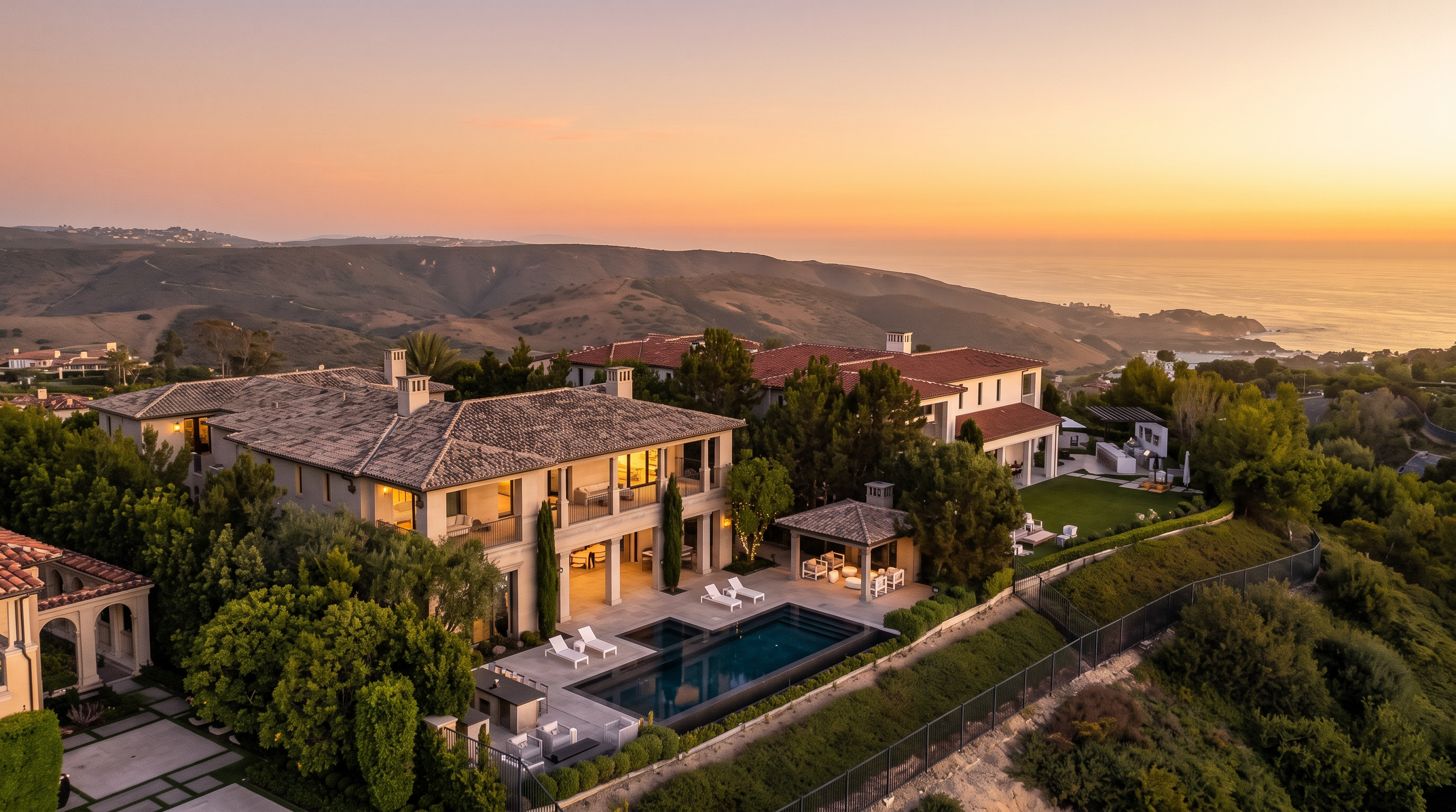 This high-angle aerial shot captures a sprawling Mediterranean-style estate at dusk, set against a backdrop of rolling hills and the distant ocean. The property features a multi-level residence with a tiled roof, a rectangular swimming pool, and a covered outdoor lounge area. The scene is bathed in the soft, purple-hued light of twilight, emphasizing the luxurious and serene atmosphere of the coastal location.