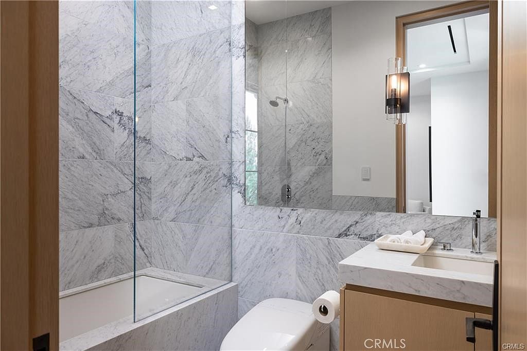 This compact, modern bathroom features striking stone-patterned wall tiles that extend into the shower area, which is enclosed by a clear glass panel. A light wood vanity with a stone countertop sits beneath a large mirror, complemented by a contemporary wall-mounted light fixture. The space is efficiently designed, incorporating a toilet and a clean, minimalist aesthetic.
