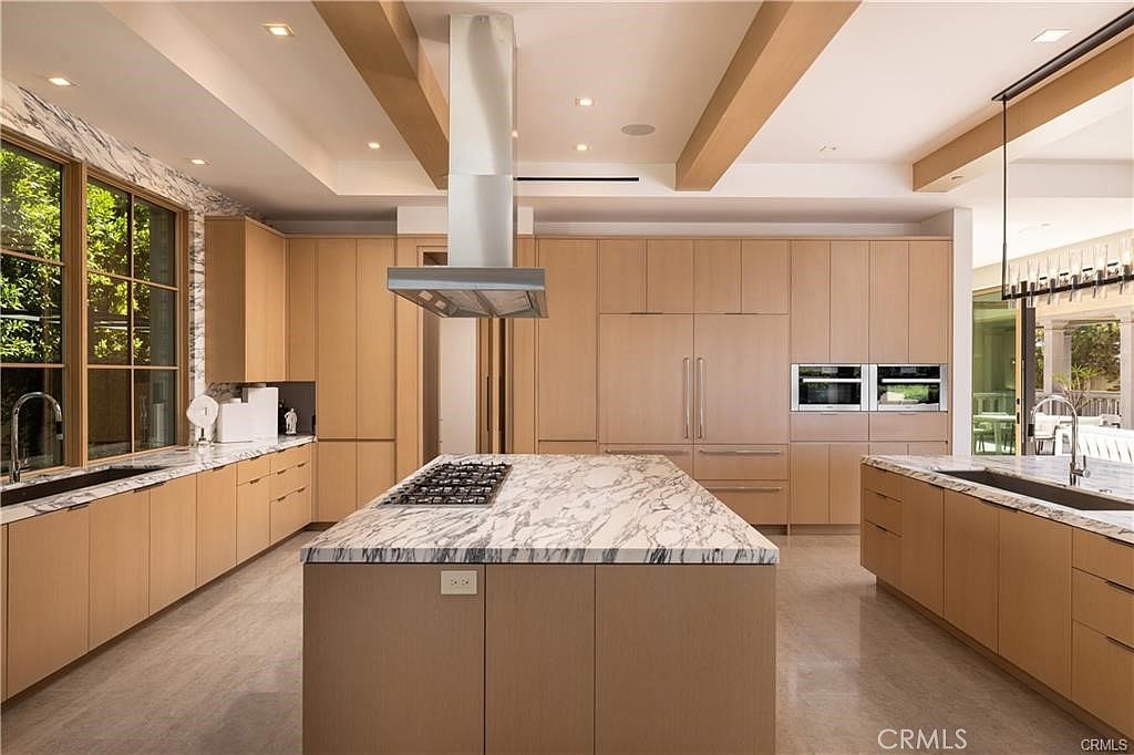 This spacious, modern kitchen features light wood cabinetry and a large central island topped with patterned stone. A stainless steel range hood hangs above the island, while integrated appliances and double wall ovens are seamlessly built into the cabinetry wall. The room is accented by exposed wooden ceiling beams and large windows that provide ample natural light, creating a bright and sophisticated atmosphere.