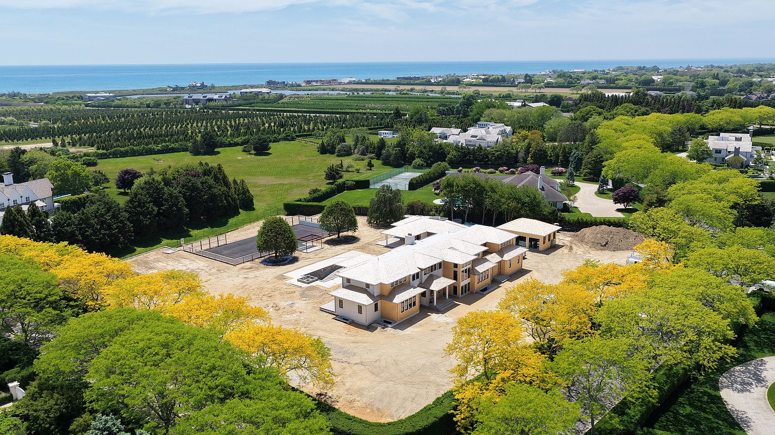 This high-angle aerial view captures a large, multi-winged residential property currently under construction, set within a lush, green landscape. The house features a light-colored roof and unfinished exterior walls, surrounded by a cleared dirt lot, a tennis court, and mature trees. In the distance, the property overlooks a vast coastal horizon, emphasizing its prime location and expansive grounds.