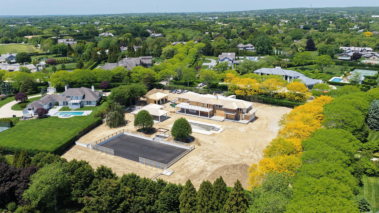 This high-angle aerial view captures a sprawling residential property currently under construction, featuring a large main residence, a detached structure, and a newly paved rectangular court. The expansive lot is surrounded by lush, mature greenery and neighboring estates, highlighting the property's significant size and potential for landscaping. The scene provides a clear perspective of the site layout, including the unfinished pool area and the surrounding manicured hedges.