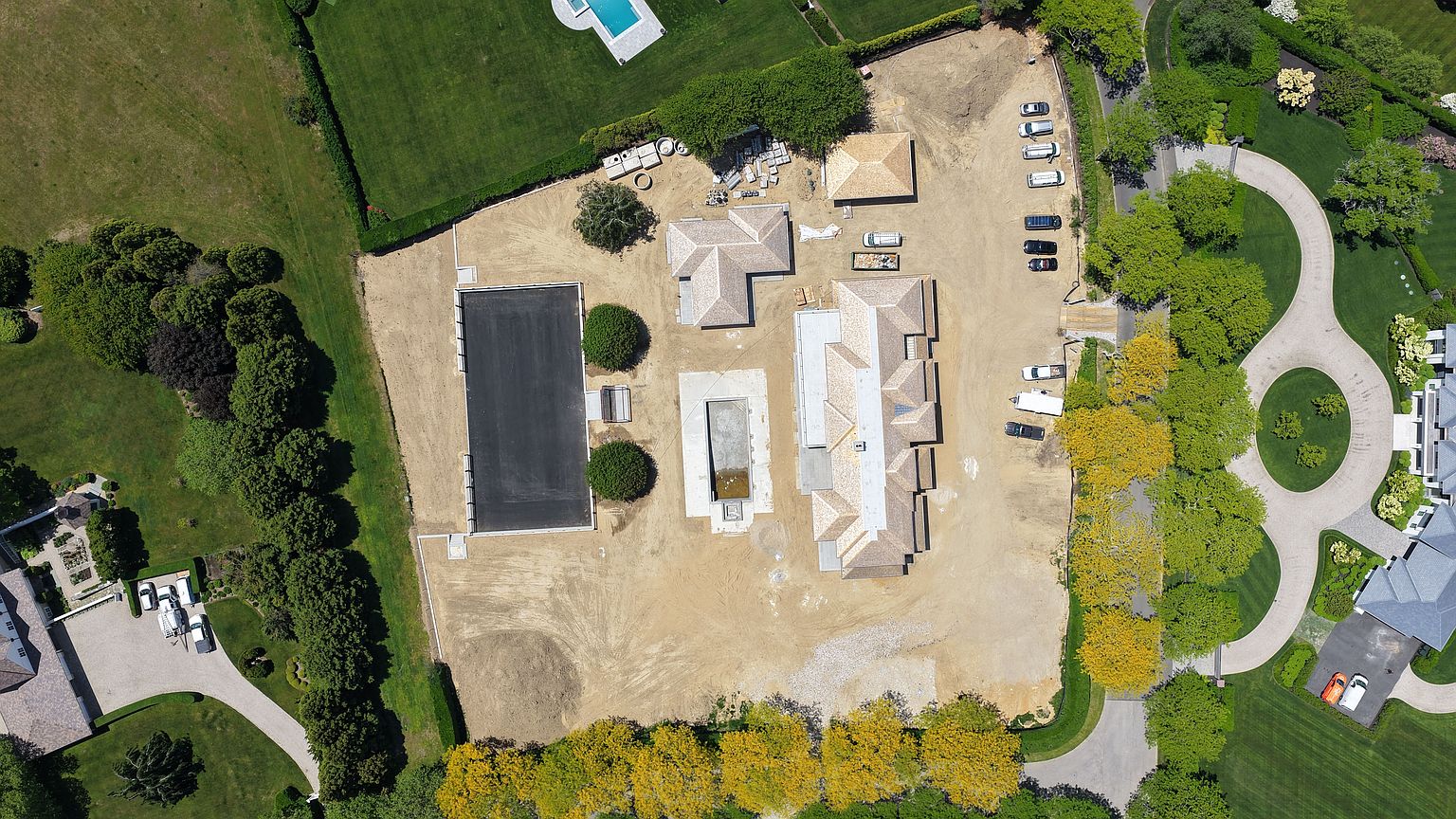 This high-angle aerial view captures a large residential construction site in the midst of development. The property features a central main house structure, a detached garage, and an in-ground pool basin, all surrounded by expansive dirt lots and mature perimeter trees. The scene provides a clear perspective of the property's layout, including the winding driveway and the surrounding landscaped grounds.