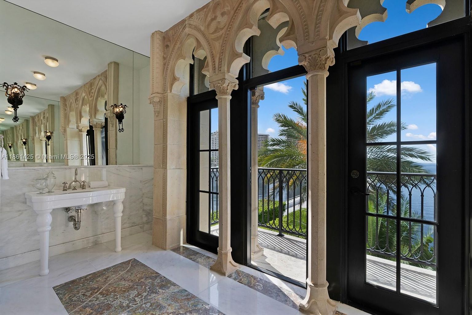 This elegant primary bathroom features a sophisticated marble vanity and a large wall-to-wall mirror, creating a bright and spacious atmosphere. The room is defined by its stunning architectural detail, showcasing ornate stone arches and columns that frame a pair of black-framed glass doors leading to a private balcony with panoramic water views. The combination of classic Mediterranean-inspired stonework and modern luxury finishes provides a refined and serene aesthetic.