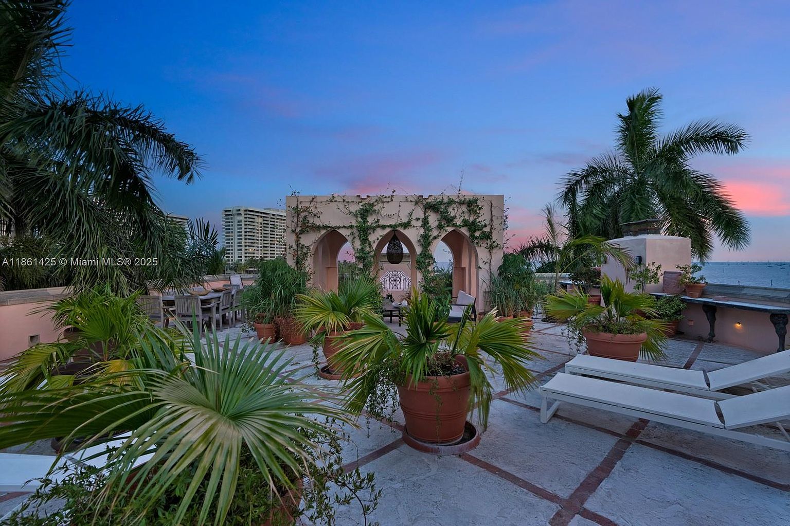 This expansive rooftop terrace offers a luxurious outdoor living space with a Mediterranean-inspired architectural structure featuring arched openings and climbing vines. The area is adorned with numerous potted palm trees and lounge chairs, set against a stunning sunset backdrop overlooking the ocean and distant city skyline. The perspective is a wide, eye-level shot that captures the serene, resort-like atmosphere of this high-end property.