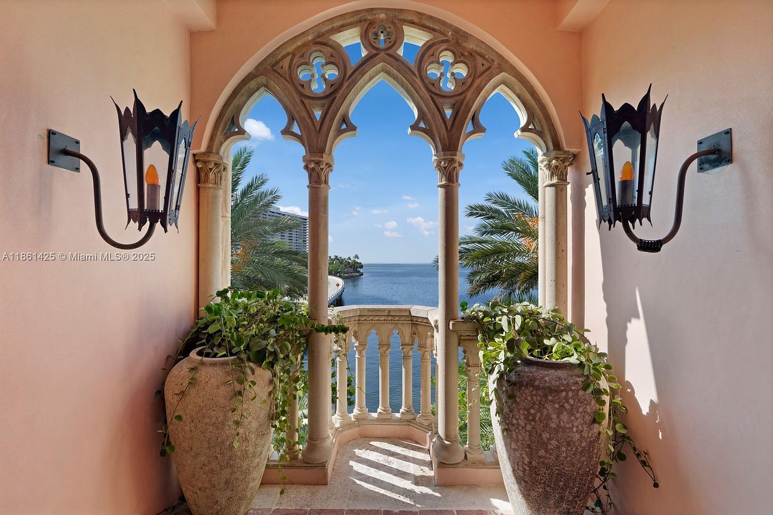This elegant balcony features intricate Gothic-inspired stone arches and columns that frame a stunning, unobstructed view of the ocean. Two large, weathered stone planters filled with lush trailing greenery flank the space, while ornate black iron wall sconces add a touch of classic sophistication. The perspective is centered and symmetrical, emphasizing the architectural craftsmanship and the serene, picturesque waterfront setting.