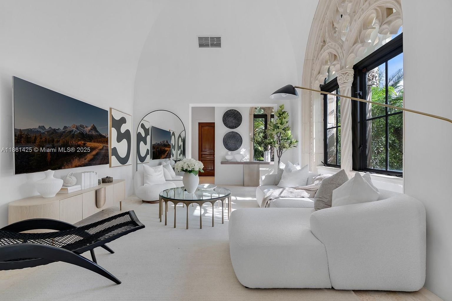 This elegant living room features a high, vaulted ceiling and a striking arched window with intricate stone detailing, creating a sophisticated and airy atmosphere. The space is furnished with plush white seating, a circular glass-top coffee table, and a large wall-mounted television, all anchored by a neutral color palette. The room blends modern luxury with architectural character, offering a bright and inviting space perfect for relaxation or entertaining.