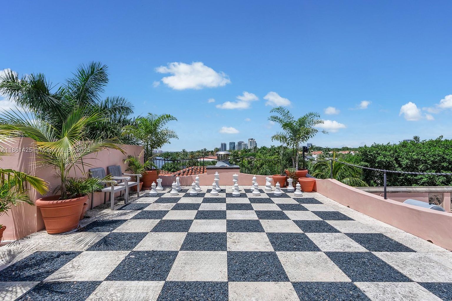 This expansive rooftop terrace features a striking black-and-white checkered floor, designed as a life-sized chessboard complete with large chess pieces. The space is framed by a low pink stucco wall, potted palm trees, and comfortable seating, offering a unique outdoor entertainment area with panoramic views of the surrounding landscape and city skyline. The perspective is a wide-angle shot from the terrace level, emphasizing the open-air atmosphere and the playful, sophisticated design.