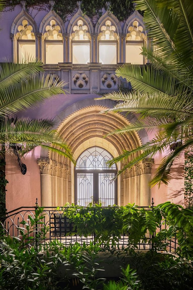 This striking entryway features ornate Gothic-inspired architecture, highlighted by a grand, multi-layered arched doorway and a row of elegant lancet windows above. The pink-hued facade is framed by lush, vibrant palm fronds and a decorative wrought-iron railing, creating a sophisticated and romantic atmosphere. The perspective is a direct, eye-level shot that emphasizes the intricate stonework and the dramatic, historic character of the home's entrance.