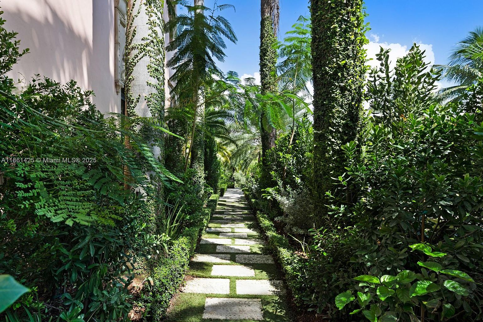 This lush, verdant garden path features a series of rectangular stone pavers set into a manicured lawn, creating a serene walkway framed by dense tropical foliage and ivy-covered tree trunks. The perspective is a low-angle, eye-level shot looking down the path, emphasizing the depth and secluded, tranquil atmosphere of the landscape. The scene is bathed in bright, natural sunlight, highlighting the rich textures of the surrounding greenery and the elegant, private nature of the outdoor space.