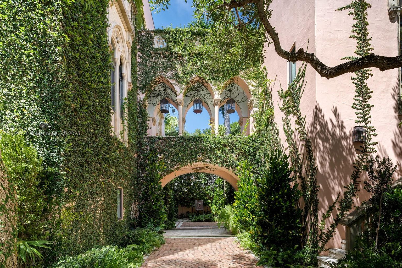 This stunning Mediterranean-style courtyard features a brick-paved walkway leading under a grand, ivy-covered stone archway. The pink stucco walls are heavily draped in lush greenery, creating a romantic and secluded atmosphere, while ornate hanging lanterns illuminate the upper loggia. The perspective is a centered, eye-level shot that emphasizes the architectural depth and the serene, garden-like quality of the space.