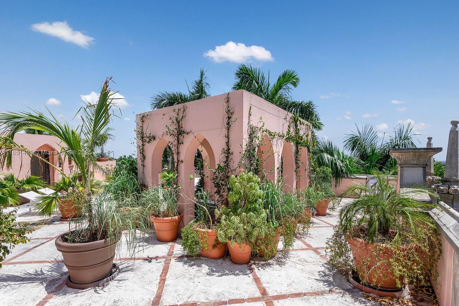 This rooftop terrace features a charming, pink-stuccoed architectural pavilion with arched openings, surrounded by lush potted plants and palm trees. The space is paved with light-colored stone tiles accented by terracotta-colored grout lines, creating a Mediterranean-inspired outdoor retreat. The perspective is eye-level, capturing the vibrant contrast between the soft pink structure, the greenery, and the bright blue sky above.