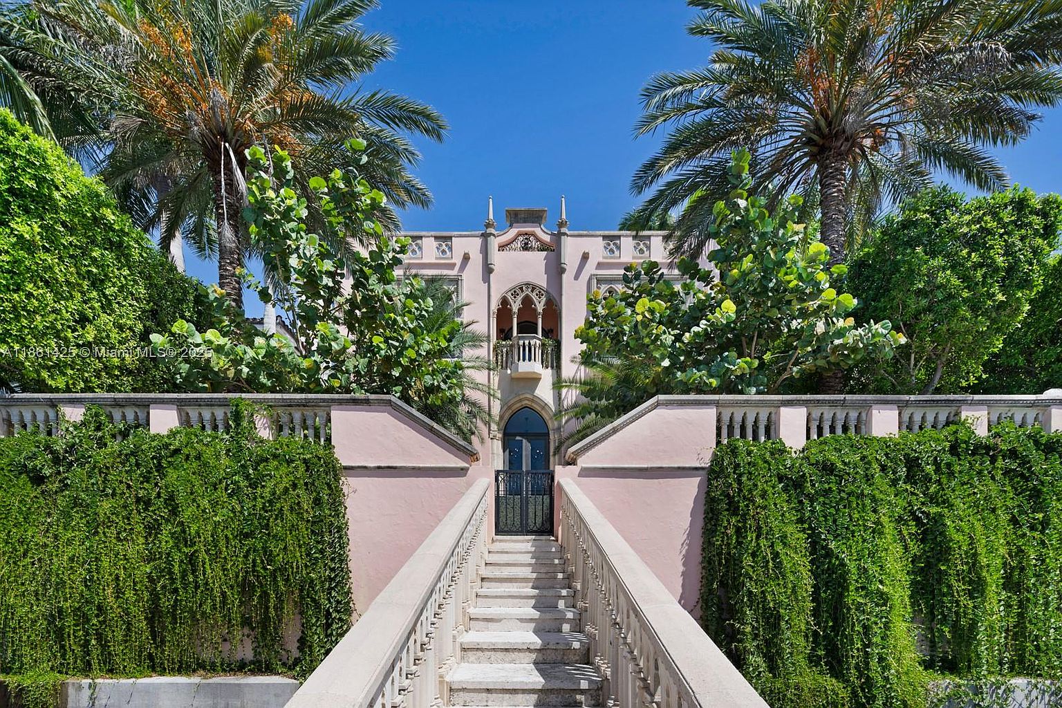 This striking Mediterranean-style mansion features a symmetrical pink facade with intricate Gothic-inspired arched windows and a prominent central balcony. A grand, wide stone staircase leads directly up to the main entrance, flanked by lush, cascading greenery and towering palm trees that frame the property. The perspective is a low-angle, centered shot that emphasizes the architectural grandeur and the imposing, elegant presence of the estate.