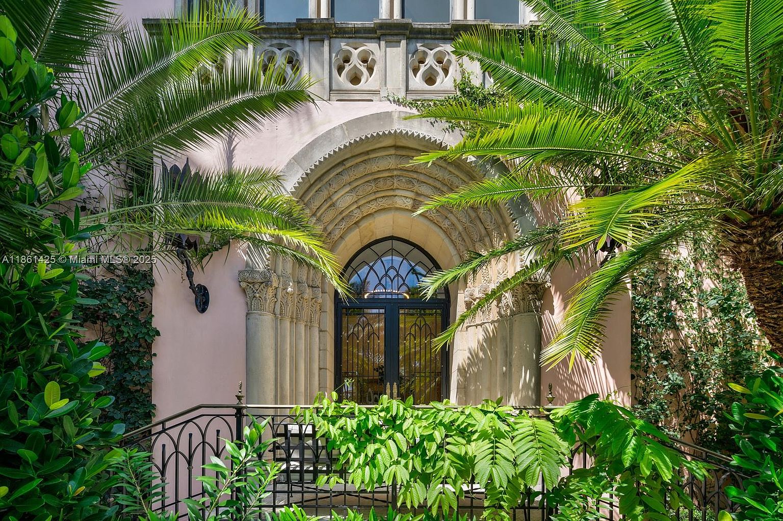 This image captures a grand, ornate entryway featuring a dramatic Gothic-style arched doorway framed by intricate stone carvings and columns. Lush, vibrant palm fronds frame the scene, creating a sense of privacy and tropical elegance, while a decorative wrought-iron railing sits in the foreground. The perspective is a direct, eye-level shot that emphasizes the architectural sophistication and historic charm of the residence.