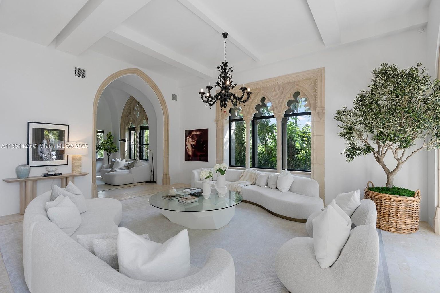This elegant living room features a sophisticated, monochromatic design with curved white sofas and a central glass-topped coffee table. The space is defined by dramatic Gothic-style arched windows and doorways, complemented by a striking black chandelier and a large potted olive tree. The overall aesthetic is one of refined luxury, blending historic architectural details with contemporary, minimalist furnishings.