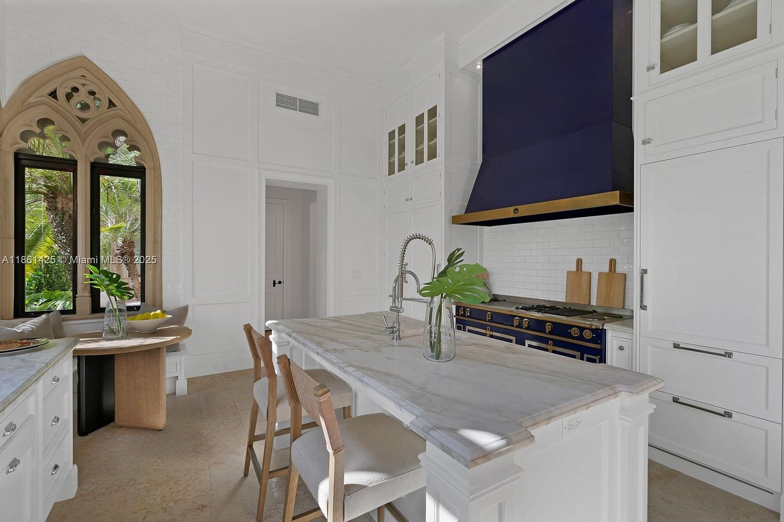 This elegant, high-end kitchen features a striking navy blue range hood and matching stove, contrasted against crisp white cabinetry and white brick walls. A large marble-topped island serves as the centerpiece, complemented by a unique arched window with gothic-inspired detailing that floods the space with natural light. The room exudes a sophisticated, timeless aesthetic with a breakfast nook area that seamlessly integrates into the refined design.