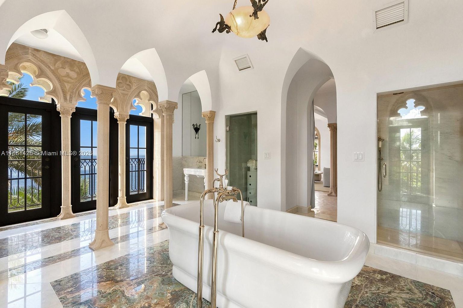 This luxurious primary bathroom features a grand, Mediterranean-inspired design with soaring arched ceilings, ornate stone columns, and a polished marble floor with intricate green and white patterns. A classic freestanding soaking tub with gold fixtures serves as the centerpiece, while floor-to-ceiling glass doors provide stunning ocean views and access to the outdoors. The space exudes opulence and tranquility, blending architectural elegance with high-end finishes.