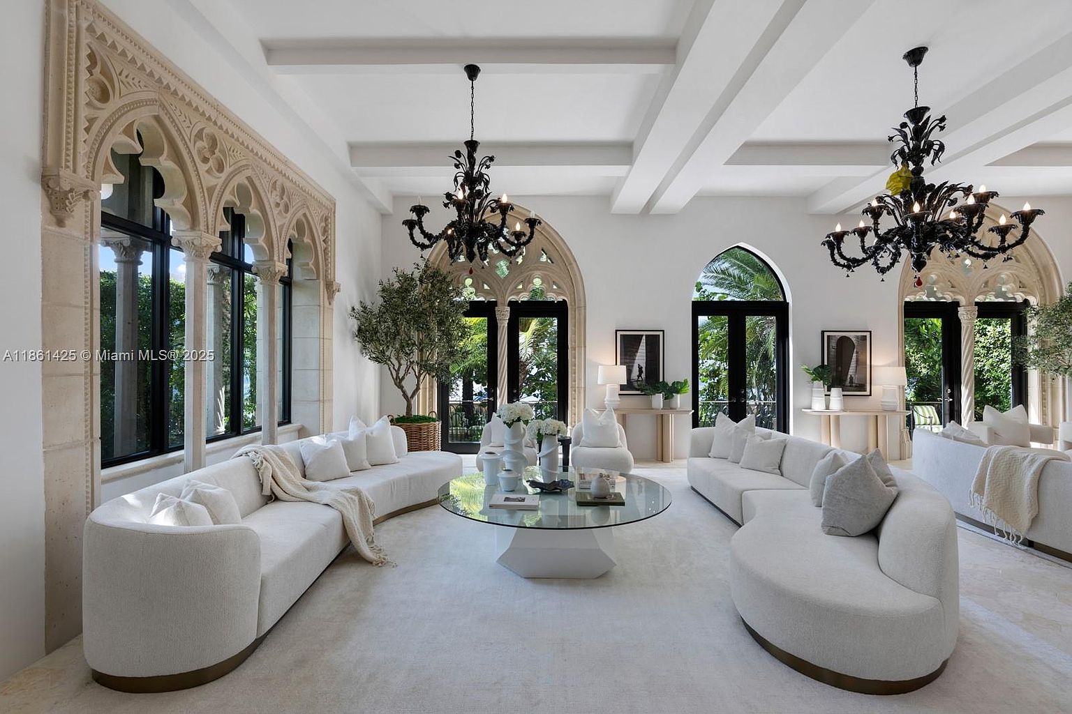 This elegant, expansive living room features a sophisticated monochromatic white palette, highlighted by dramatic Gothic-inspired arched windows and ornate stone detailing. The space is furnished with two large, curved white sofas arranged around a central glass-topped coffee table, all illuminated by two striking black wrought-iron chandeliers. The room offers a bright, airy atmosphere with a seamless blend of classical architectural elements and contemporary luxury design.