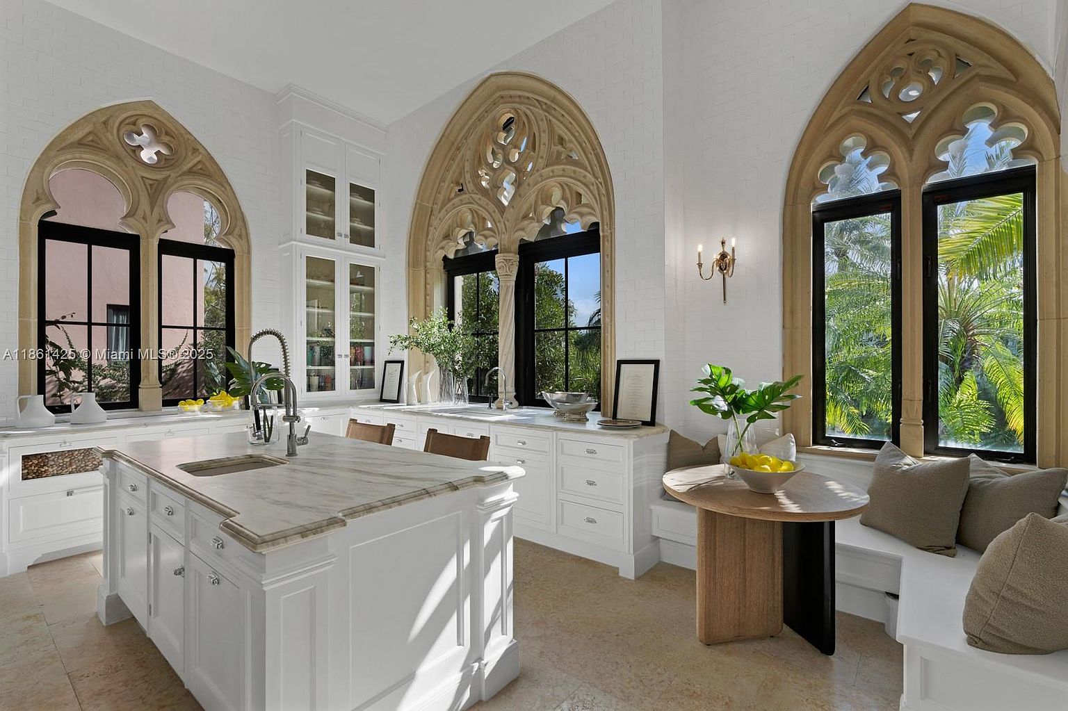 This elegant, light-filled kitchen features a sophisticated blend of classic Gothic architectural elements and modern luxury. The space is anchored by a large marble-topped island and custom white cabinetry, complemented by unique arched stone-framed windows that offer lush garden views. A cozy breakfast nook with built-in bench seating adds a warm, inviting touch to this bright and airy culinary space.