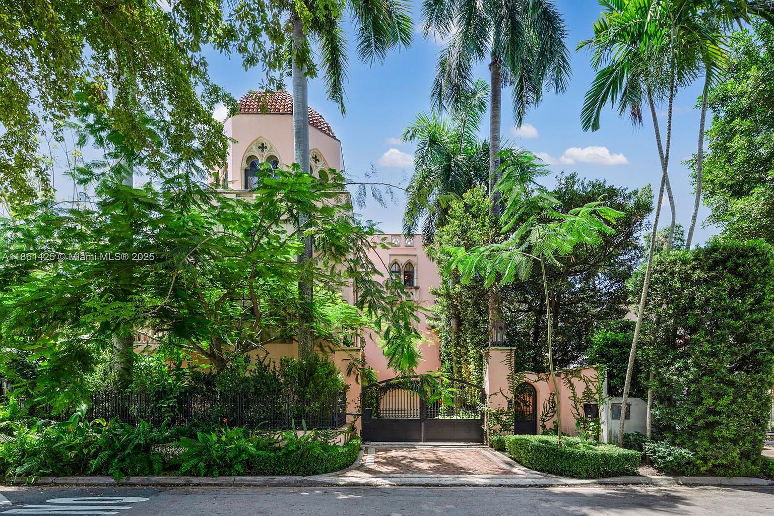 This striking Mediterranean-style estate features a distinctive pink stucco facade, a prominent domed tower, and intricate arched windows. The property is framed by lush, mature tropical landscaping and tall palm trees, with a gated brick driveway providing a grand, private entrance. The perspective is a straight-on eye-level shot that emphasizes the architectural elegance and secluded, upscale nature of the residence.
