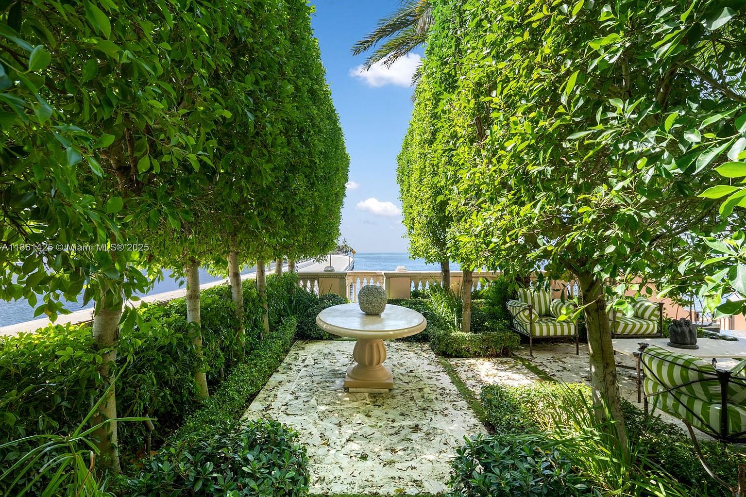 This elegant, manicured garden terrace offers a serene outdoor retreat framed by tall, perfectly trimmed hedges that create a natural corridor leading toward the water. A central stone pedestal table serves as a focal point, complemented by striped outdoor seating nestled among lush greenery and stone pavers. The perspective captures a tranquil, resort-like atmosphere with a clear view of the ocean beyond the stone balustrade.