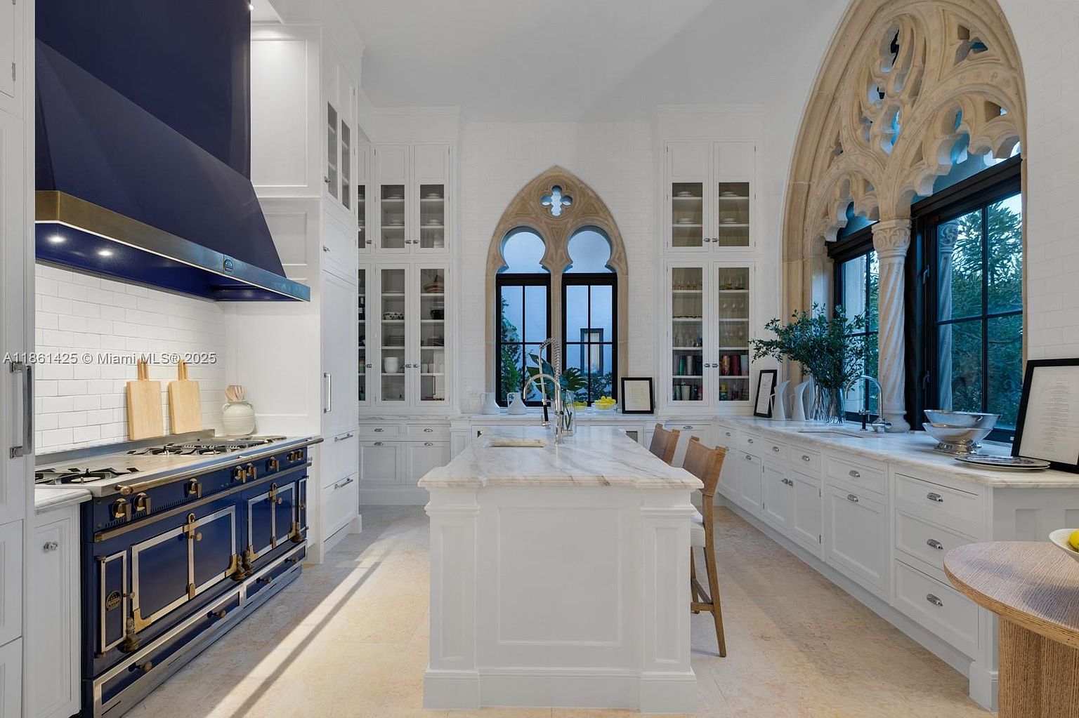 This elegant, high-end kitchen features a striking navy blue range and matching hood, contrasted against crisp white cabinetry and marble countertops. The space is defined by its unique Gothic-inspired arched windows with intricate stone detailing, creating a sophisticated and timeless atmosphere. A central island with bar seating serves as the focal point, perfectly blending classic architectural elements with modern luxury.
