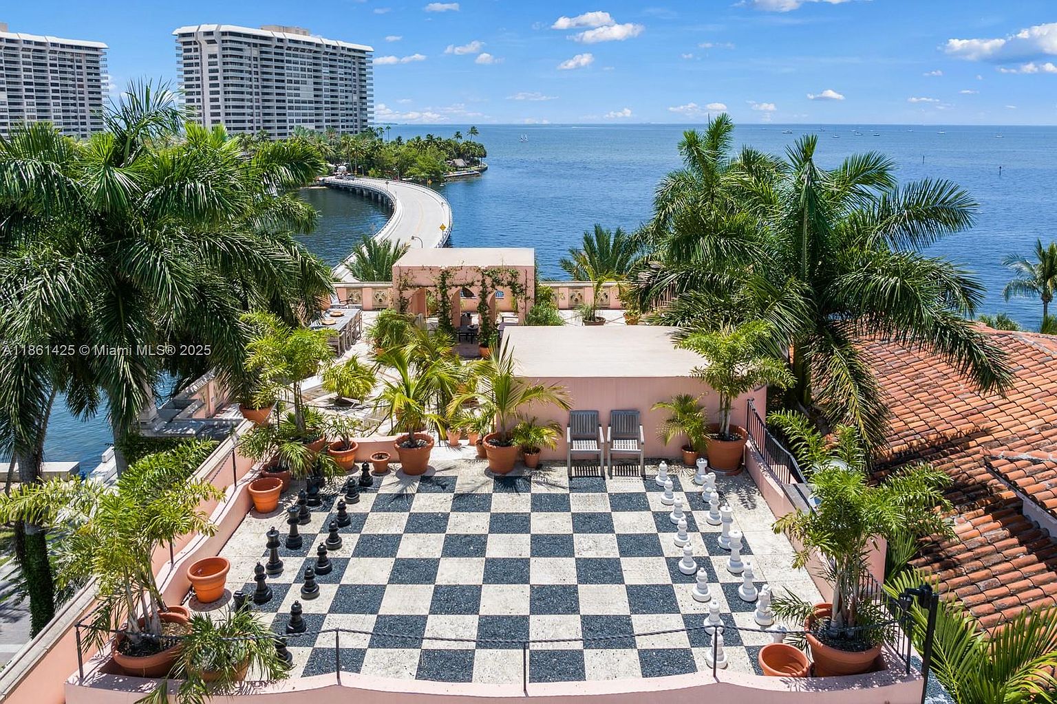 This elevated rooftop terrace features a unique, large-scale outdoor chessboard as its centerpiece, surrounded by lush potted palm trees and terracotta planters. The space offers a stunning, panoramic view of the bay, a curved bridge, and distant high-rise buildings, creating a luxurious and serene atmosphere. The perspective is captured from an elevated vantage point, emphasizing the expansive outdoor living area and its picturesque coastal backdrop.