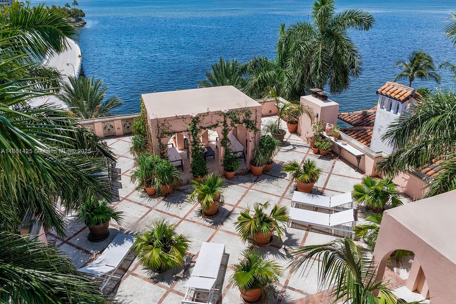 This high-angle aerial view captures a spacious, Mediterranean-style rooftop terrace overlooking a serene blue bay. The area features a central pergola draped in climbing vines, multiple potted palm trees, and several white lounge chairs arranged for relaxation. The stone-tiled deck, accented with terracotta-colored grout lines, creates a warm, inviting atmosphere perfect for outdoor entertaining.