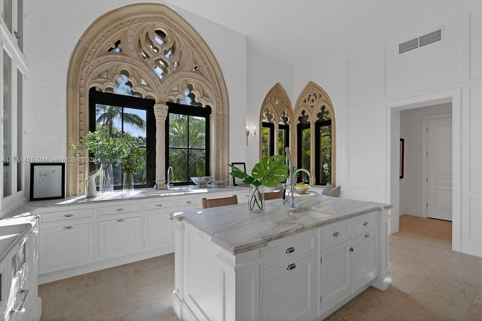 This elegant, high-end kitchen features a bright, all-white aesthetic with a large marble-topped island and custom cabinetry. The space is defined by its striking Gothic-inspired arched windows with intricate stone tracery, which flood the room with natural light and provide a view of the lush greenery outside. The combination of classic architectural details and modern luxury finishes creates a sophisticated and airy atmosphere.