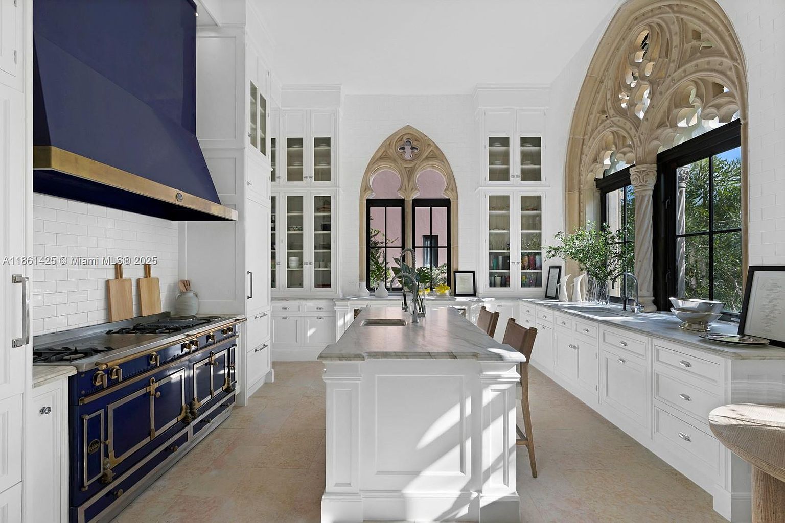 This elegant, high-end kitchen features a striking navy blue range and matching hood, contrasted by crisp white cabinetry and marble countertops. The space is defined by its unique Gothic-inspired arched windows that provide a dramatic architectural focal point. A central island with seating creates a functional workspace, while the overall aesthetic blends classic luxury with sophisticated design.