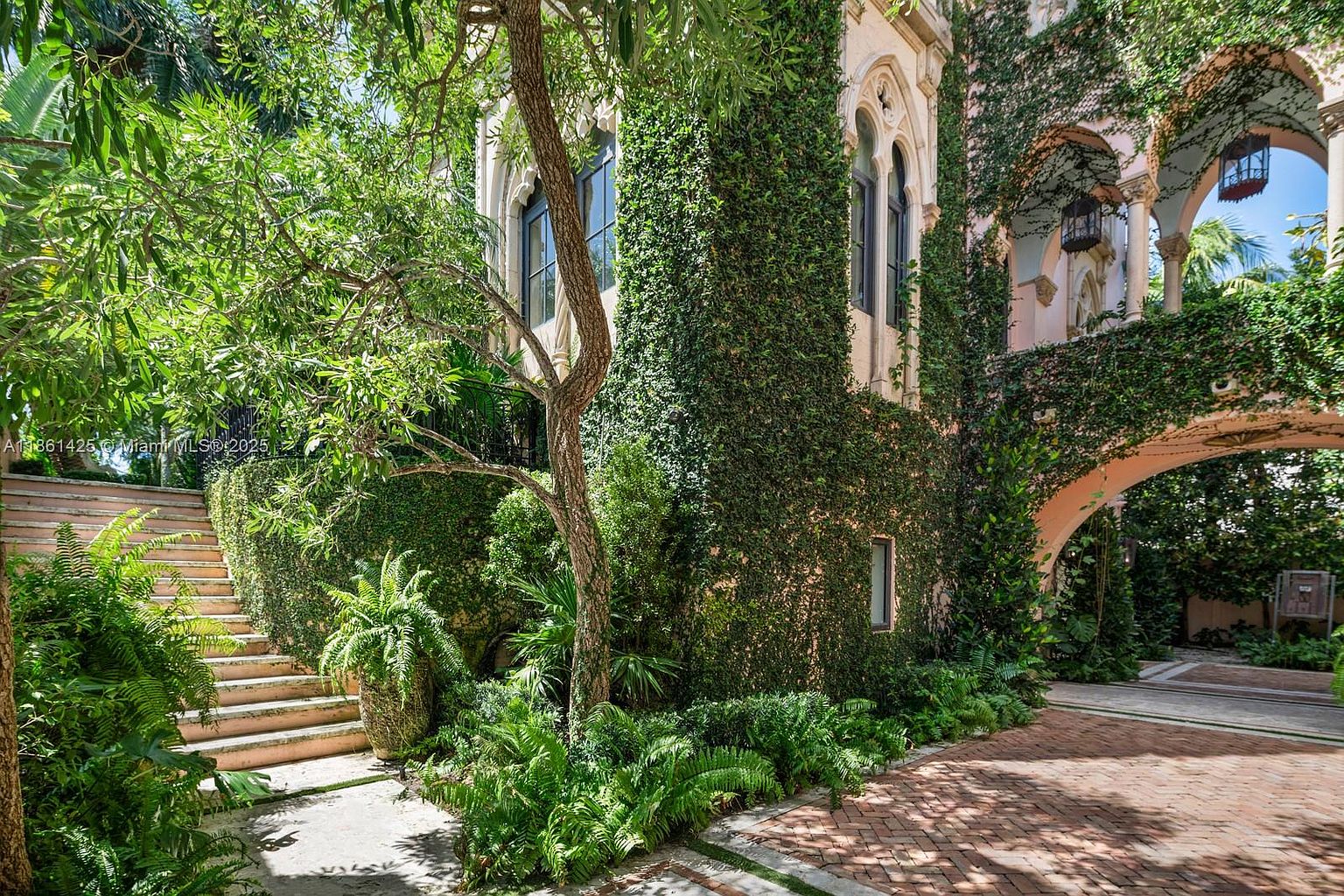 This enchanting exterior scene showcases a Mediterranean-style villa draped in lush climbing ivy, featuring elegant arched windows and a stone-paved courtyard. A set of stone stairs leads up through a verdant garden filled with ferns and tropical foliage, creating a serene and historic atmosphere. The perspective captures the interplay between the architectural stonework and the vibrant greenery, evoking a sense of timeless luxury and privacy.