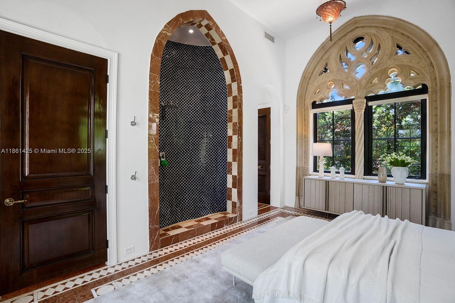 This elegant primary bedroom features a striking arched stone entryway leading to a tiled shower, complemented by a dramatic, ornate Gothic-style arched window that serves as a focal point. The room is furnished with a plush bed and a sleek, low-profile console, blending classic architectural details with modern interior design. The perspective is a wide-angle shot from the foot of the bed, capturing the sophisticated contrast between the dark wood door, the intricate stone archway, and the bright, airy window area.