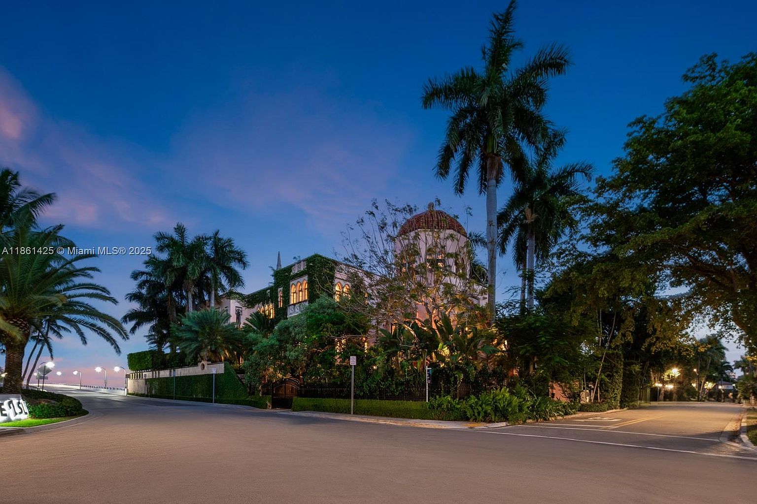 This stunning Mediterranean-style estate is captured at twilight, showcasing its grand architecture, including a prominent central dome and lush ivy-covered walls. The property is framed by mature palm trees and manicured landscaping, set against a vibrant, deep blue evening sky. The perspective is from the street level, emphasizing the home's imposing presence and elegant curb appeal.