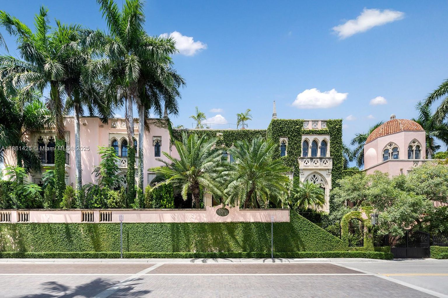 This grand, Mediterranean-style estate features a striking pink facade adorned with lush climbing ivy and intricate architectural details. The property is framed by towering palm trees and a manicured hedge wall, creating a sense of privacy and opulence. The perspective is a wide, eye-level shot from the street, emphasizing the impressive scale and historic charm of the residence.