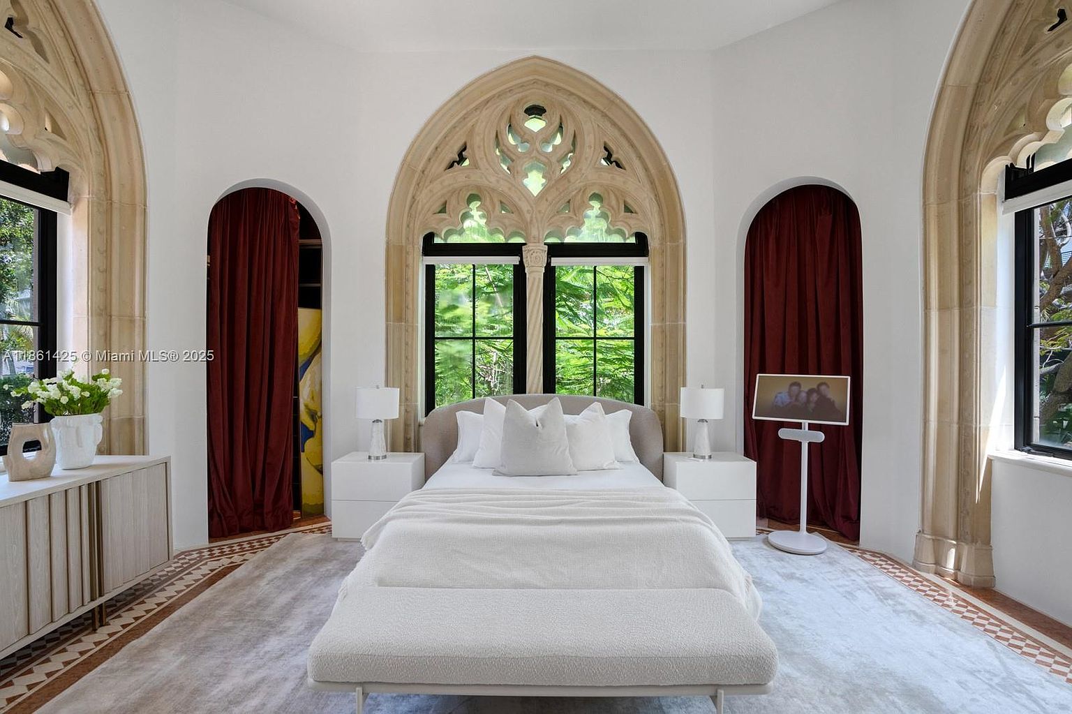 This elegant primary bedroom features a sophisticated, neutral color palette centered around a plush bed with white linens and a matching bench. The room is defined by its striking Gothic-inspired arched stone window frames and intricate architectural detailing, which frame views of lush greenery outside. A symmetrical layout with white nightstands and lamps creates a serene, high-end atmosphere, complemented by rich, deep red curtains that add a touch of dramatic contrast.