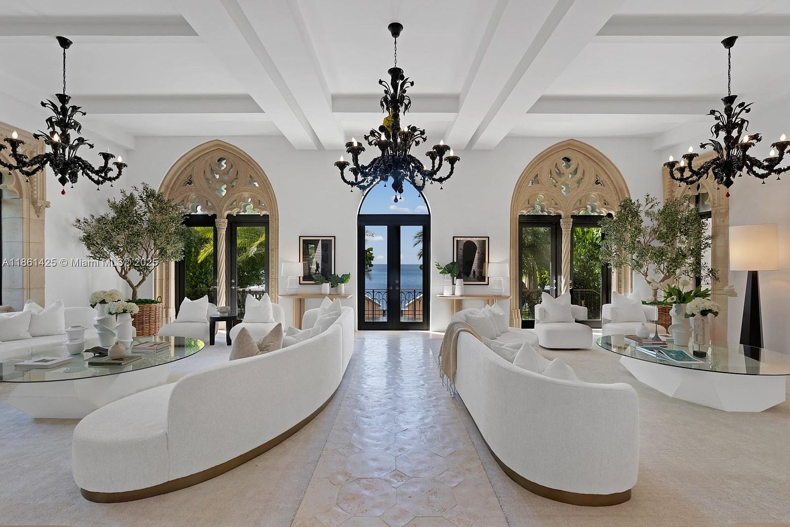 This grand, symmetrical living room exudes Mediterranean elegance with its soaring coffered ceilings, arched gothic-style windows, and ornate black chandeliers. The space is anchored by two large, curved white sofas facing each other, centered around glass-topped coffee tables and a light-toned stone floor. The room offers a stunning, unobstructed view of the ocean through the central doorway, creating a sophisticated and airy atmosphere.