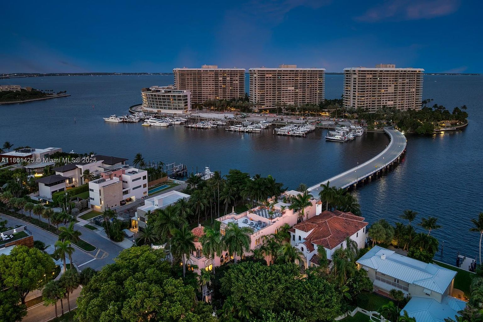 This high-angle aerial view captures a luxurious waterfront estate in a tropical setting, featuring a Mediterranean-style mansion with a red-tiled roof and lush landscaping. A private bridge connects the property to a nearby marina filled with yachts, set against the backdrop of large residential high-rises and the expansive bay. The scene is captured during the twilight hour, creating a serene and sophisticated atmosphere that highlights the exclusive nature of the location.