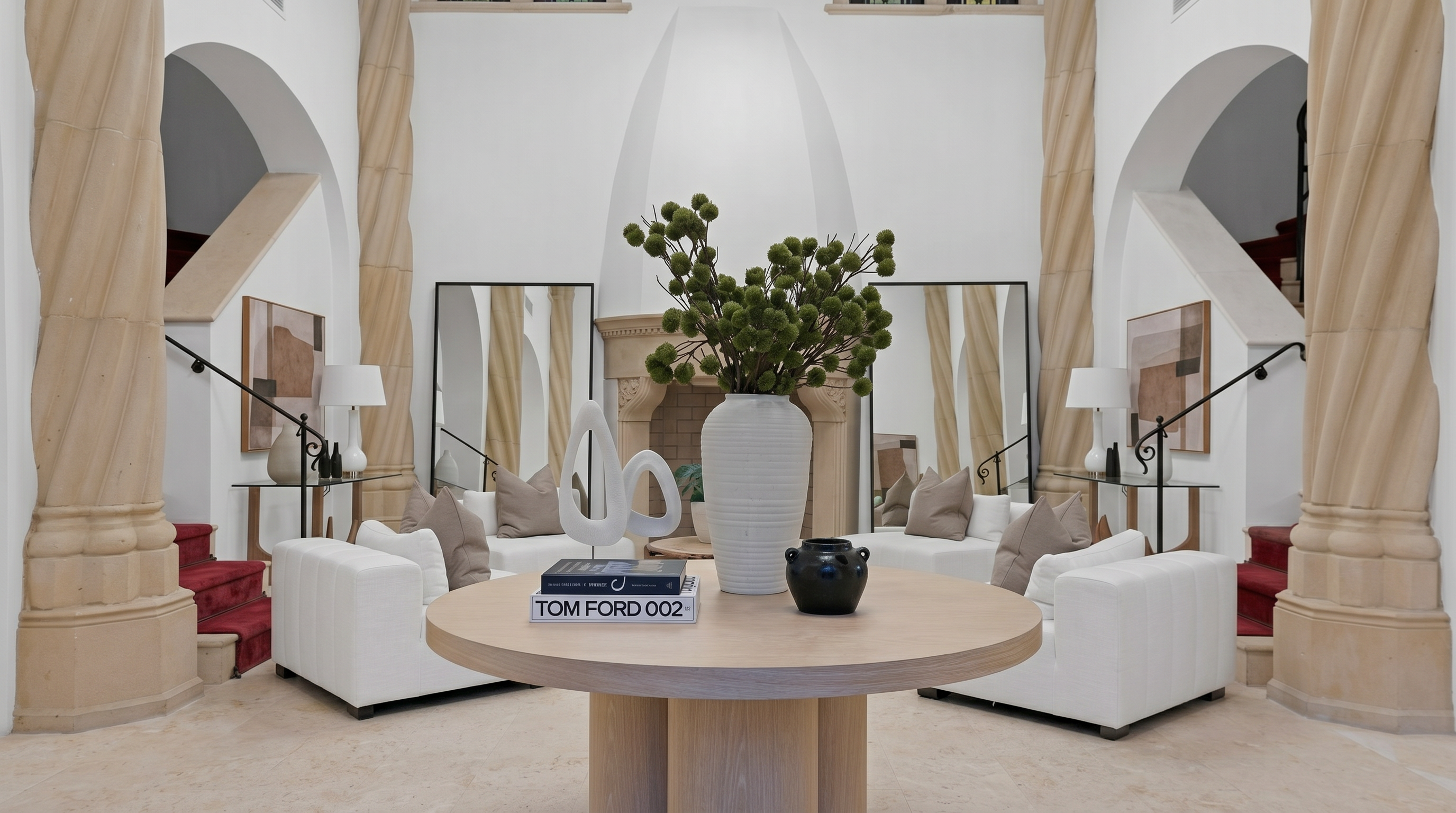 This elegant and symmetrical living room features a sophisticated, neutral color palette with white sofas, tan throw pillows, and light wood textures. The space is anchored by a large circular wooden accent table, grand stone fireplace, and striking architectural columns that frame dual staircases. The high ceilings and mirrored accents enhance the airy, luxurious, and refined atmosphere of the residence.