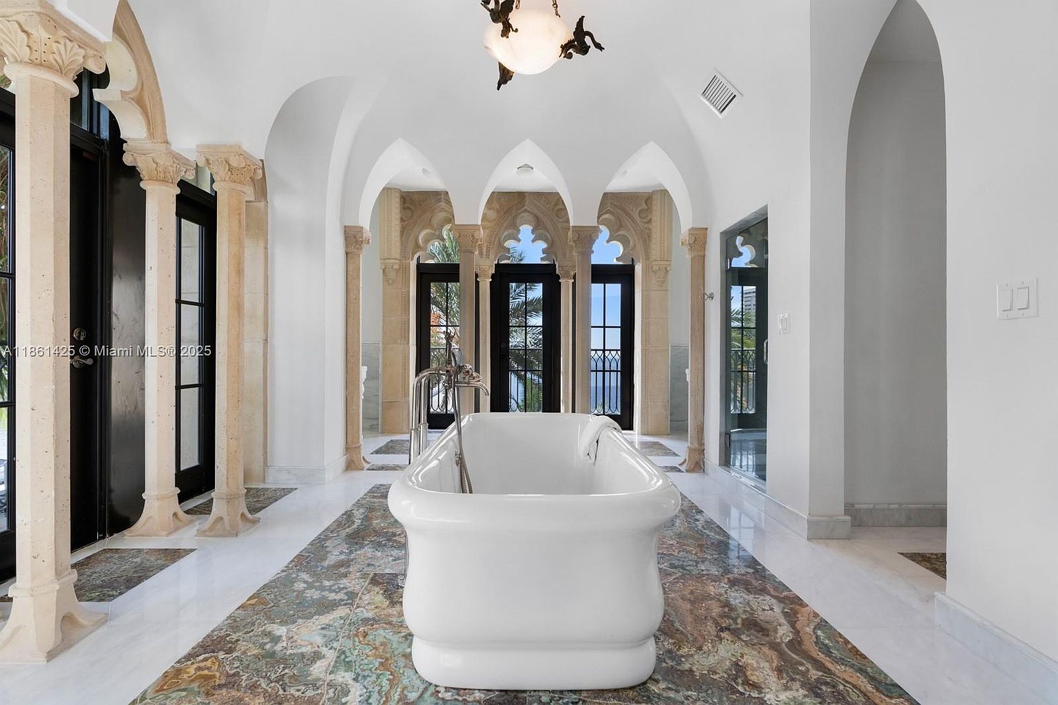 This luxurious primary bathroom features a grand, freestanding white soaking tub as its centerpiece, set against a backdrop of ornate stone columns and arched doorways. The room is defined by its high, vaulted ceilings and a striking floor inlay of multi-colored marble that contrasts beautifully with the surrounding white marble flooring. The space exudes an elegant, Mediterranean-inspired aesthetic with expansive windows that provide a bright, airy atmosphere and scenic views.