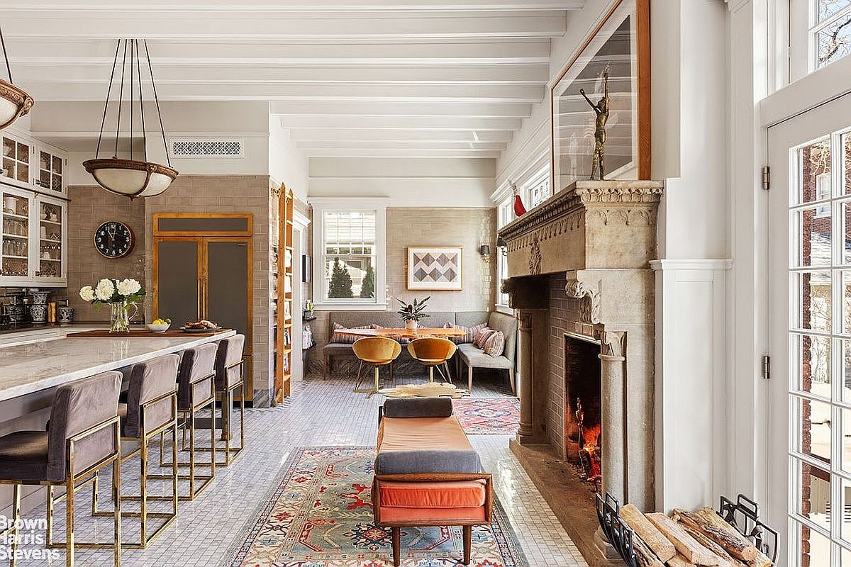 This interior shot showcases a stylish living room with a blend of modern and classic elements. The room features a large stone fireplace, a built-in banquette seating area, and a kitchen island with bar stools. The space is well-lit and decorated with rugs and artwork, creating a warm and inviting atmosphere.