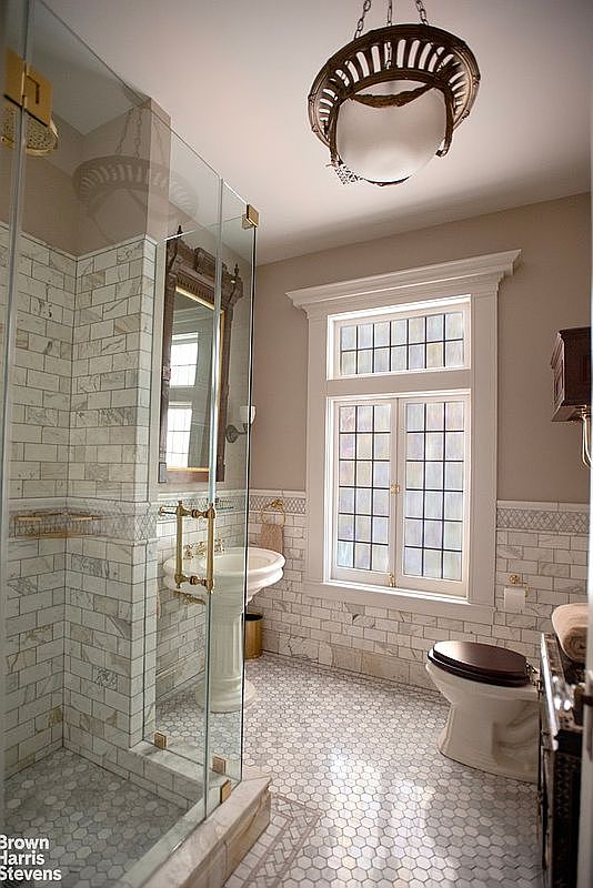 This is a primary bathroom featuring a glass-enclosed shower with marble subway tile, a pedestal sink with brass fixtures, and a toilet with a dark wood seat. The floor is covered in a small hexagonal tile pattern, and a window provides natural light. The overall style is classic and elegant.