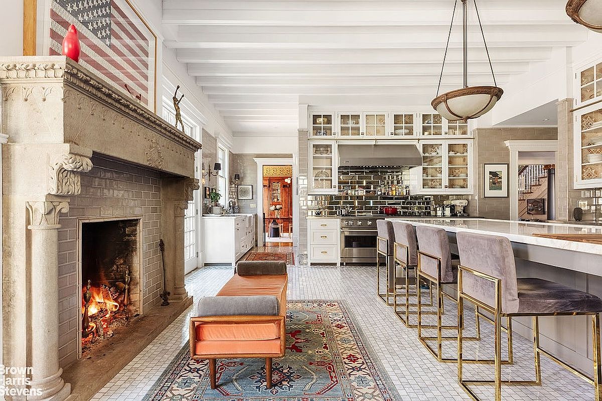 This is a spacious, high-end kitchen featuring a large fireplace, a long island with bar seating, and custom cabinetry with glass fronts. The kitchen is well-lit with a combination of natural light and pendant lighting, and the overall design is a blend of traditional and modern elements. The perspective is from the side, showcasing the length of the kitchen and the various features.
