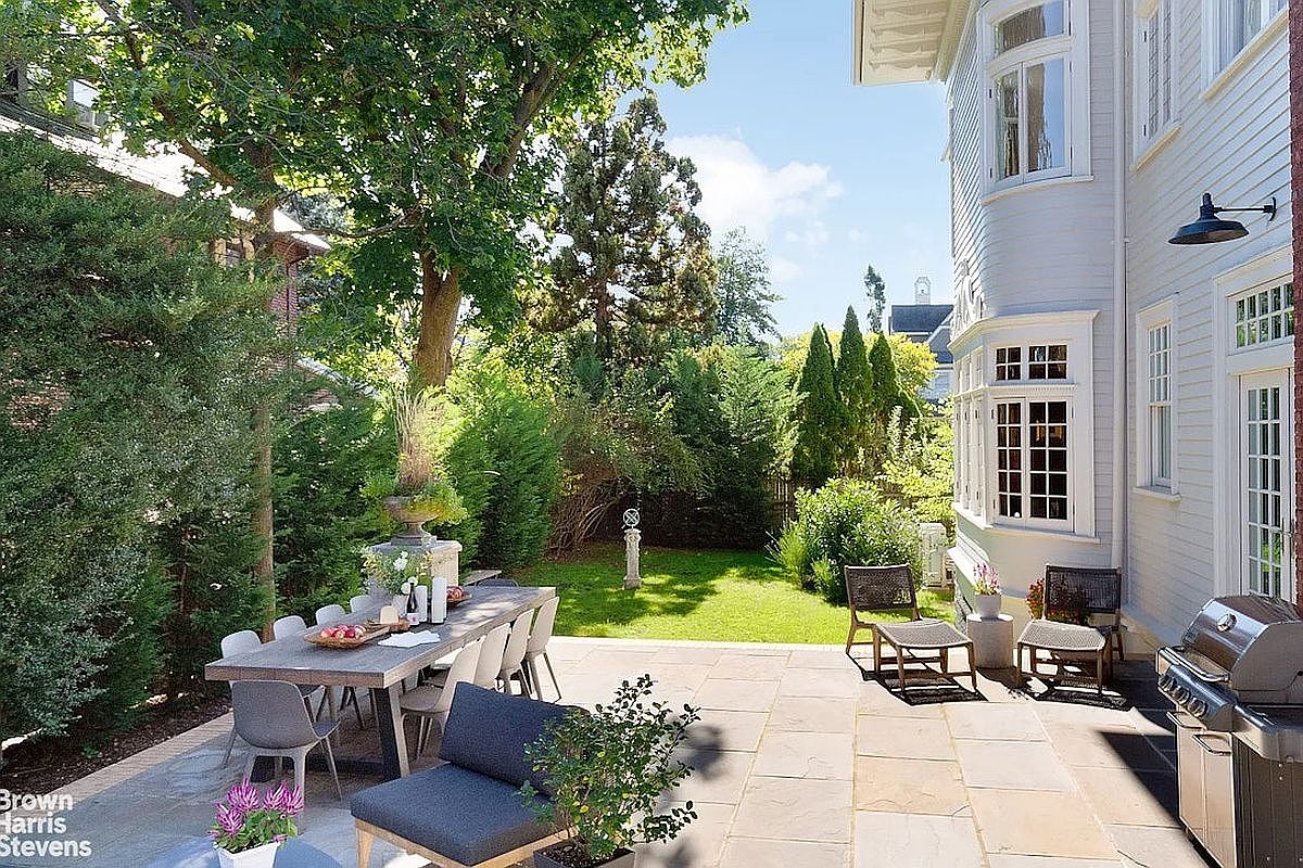 This image showcases a beautifully designed patio area, perfect for outdoor entertaining. A long dining table with modern chairs sits on a stone patio, adjacent to a lush green lawn and mature trees providing privacy. The patio also features comfortable lounge seating and a grill, creating an inviting and functional outdoor living space next to the house.