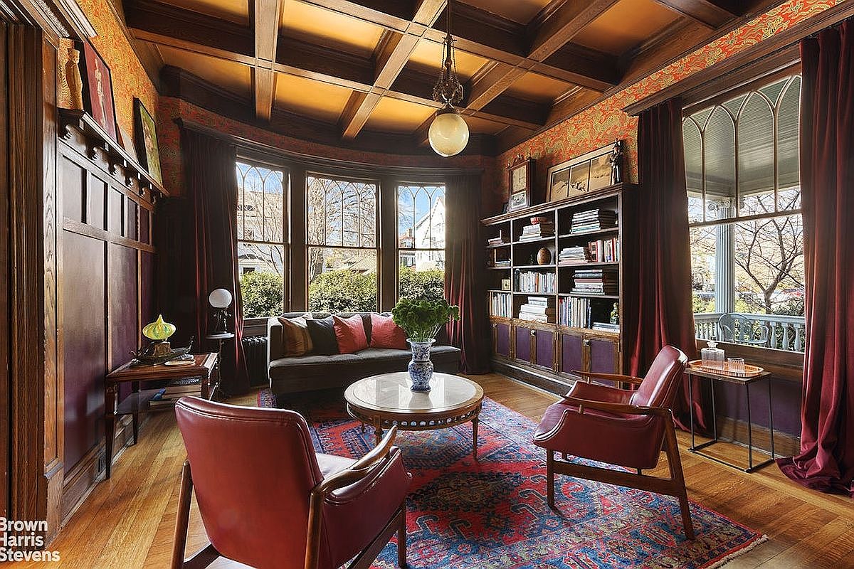 This is a beautifully designed living room featuring a coffered ceiling, a bay window with a view of the outdoors, and a built-in bookcase. The room is furnished with a sofa, two armchairs, and a round coffee table, all arranged on an ornate rug. The overall style is classic and elegant, creating a warm and inviting atmosphere.