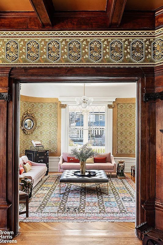 This interior shot showcases a beautifully decorated living room, viewed through an ornate wooden doorway. The room features a patterned rug, two pink sofas, and a tufted ottoman, complemented by a large window that provides natural light. The walls are adorned with patterned wallpaper and a decorative mirror, creating a warm and inviting atmosphere.