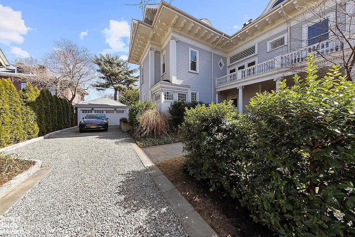 This image showcases the front exterior of a stately home with a light blue facade and white trim. The property features a gravel driveway leading to a detached garage, lush landscaping, and architectural details such as columns and a balcony. The overall impression is one of classic elegance and curb appeal.