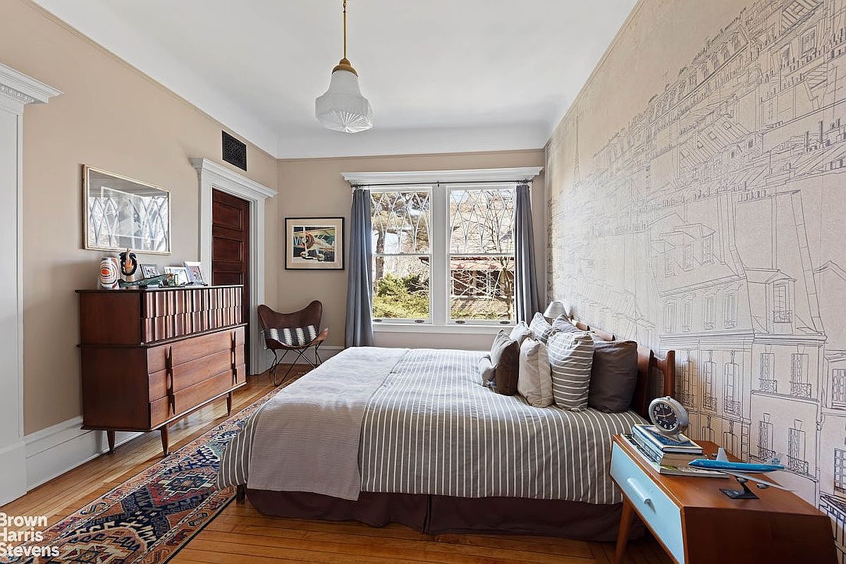 This is a cozy primary bedroom featuring a queen-sized bed with striped bedding and multiple pillows. A mid-century modern dresser and a blue nightstand flank the bed, while a unique cityscape mural adorns the wall. The room is well-lit by natural light from the windows, creating a warm and inviting atmosphere.